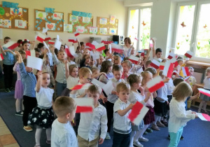 Dzieci stoją na sali, śpiewają piosenkę i machają samodzielnie wykonanymi flagami.