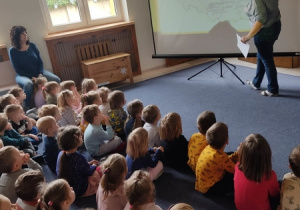 Dzieci siedzące na dywanie i słuchają nauczycielki i oglądają wyświetloną na rzutniku mapę