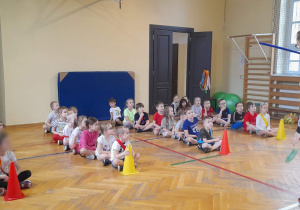 Dzieci siedzą w trzech rzędach na sali gimnastycznej